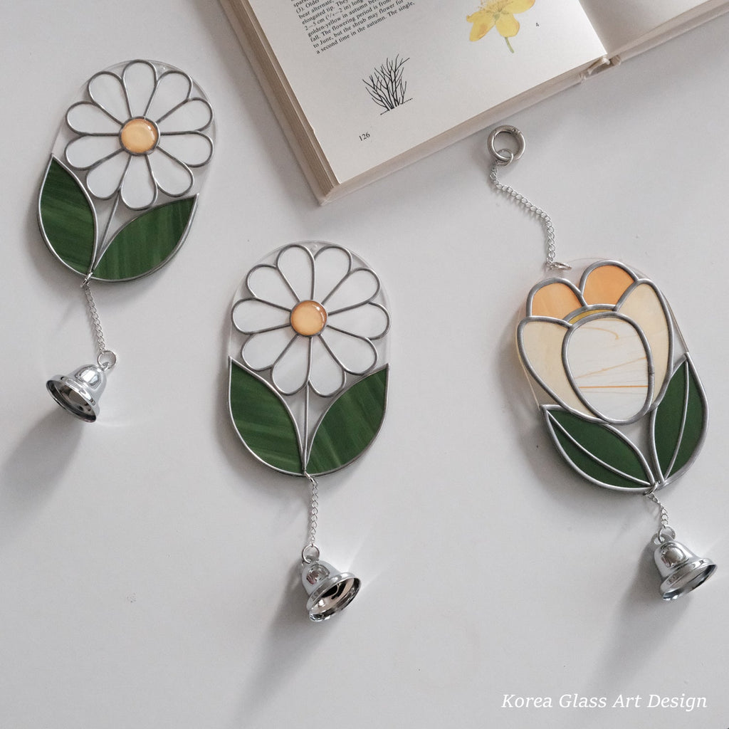 Three stained glass wall art pieces with hanging bells are arranged on a light surface beside an open book. Two pieces depict white daisies with green leaves and yellow centers, and one depicts a tulip in cream, yellow, and green with silver metal outlines and chains.