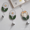 Three stained glass wall art pieces with hanging bells are arranged on a light surface beside an open book. Two pieces depict white daisies with green leaves and yellow centers, and one depicts a tulip in cream, yellow, and green with silver metal outlines and chains.