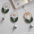 Three stained glass wall art pieces with hanging bells are arranged on a light surface beside an open book. Two pieces depict white daisies with green leaves and yellow centers, and one depicts a tulip in cream, yellow, and green with silver metal outlines and chains.