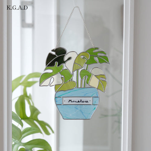 A stained glass suncatcher shaped like a potted Monstera plant, hanging in front of a window. The design features green and white monstera leaves in a light blue pot with a silver metal chain.